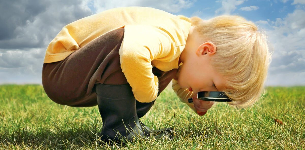 micromanage - A young boy with blonde hair wearing a long sleeve yellow shirt and brown pants tucked into black rain boots is crouched and bent over with a magnifying glass pressed to his eyes intently looking at something in the grass
