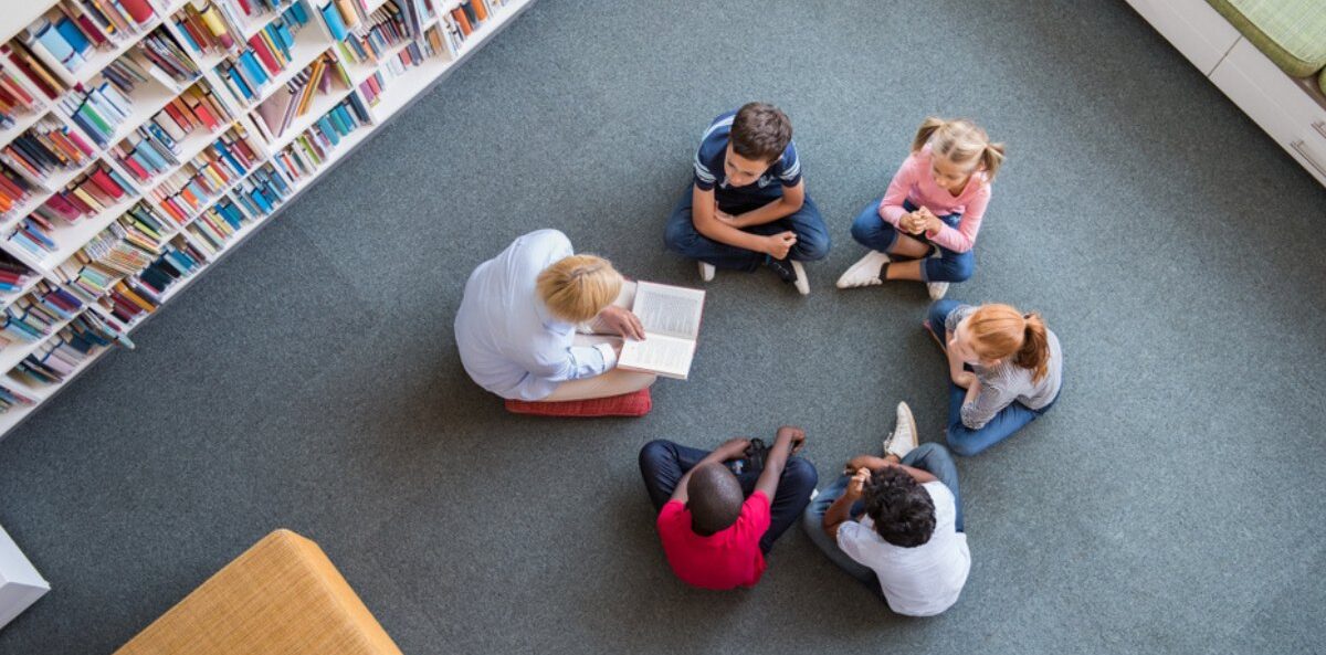 nonprofit storytelling - top view of 5 kids with different skin tones sitting in a circle facing an older person reading a book on the floor of a library