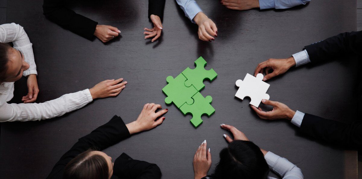 board engagement - a top view of 6 people around a black flat surface with their hands extended towards the center. In the middle are three large green connected puzzle pieces, and one of the sets of hands is extending a white puzzle piece to add to the connection.