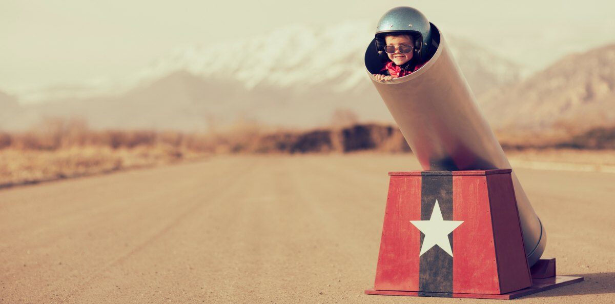 leadership conversations - A young child with light skin wearing a large, dark, shiny helmet and sunglasses is stuffed into a barrel of a pretend cannon propped up by a red blog with a navy vertical strip and large white star in the middle. The cannon is in the middle of an empty plane runway with mountains in the background.