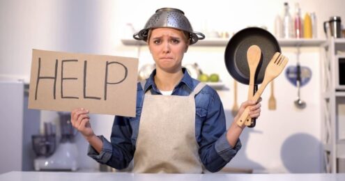Fundraising Lunch - A white female presenting person in a jean shirt and canvas apron with a metal strainer on their head like a hat while standing in a kitchen. In one hand they are holding a cardboard sign with the word help written on it, and in the other hand are wooden utensils and a cast iron pan.