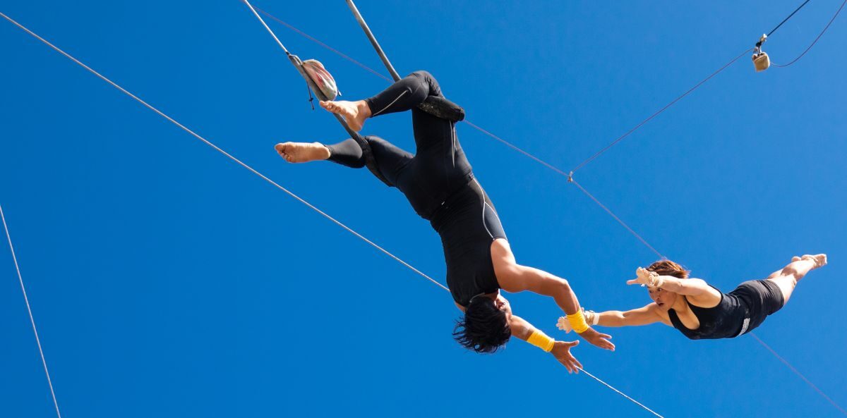 nonprofit administrative support - Two acrobat performers flying through the air. One has their arms outstretched reaching for the other mid-jump and the second is upside down wrapped around supports with their arms reached out ready to catch the first.