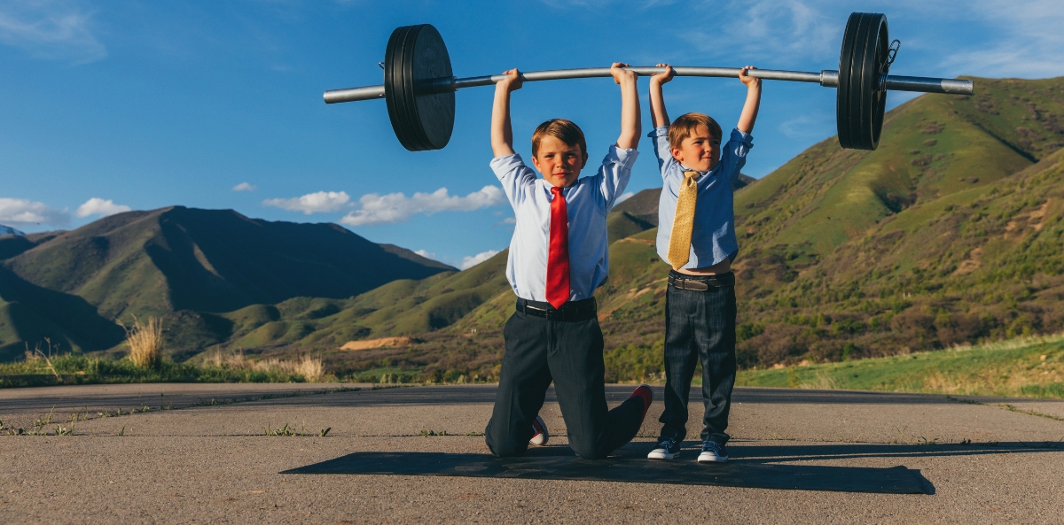 two children dressed in business clothes with ties holding up a big barbell above their heads with green mountains in the background.
