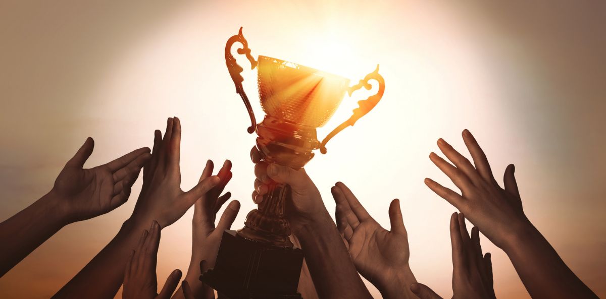 Post header image shows a bunch of hands raising up towards a trophy signifying that Executive Directors can crush your Annual Review in 10 Steps For Executive Directors